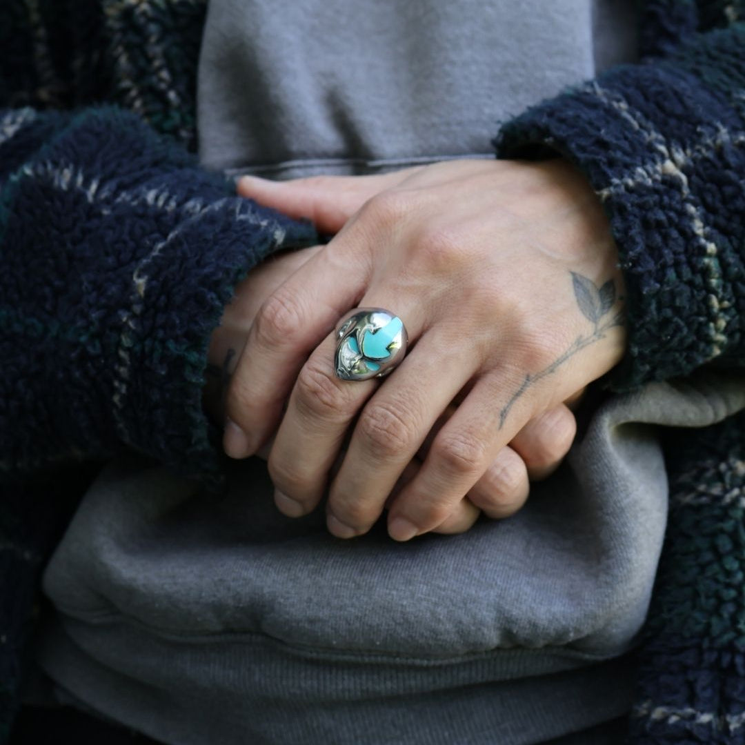 A close-up of a person's hands clasped in front of a gray sweatshirt, wearing a dark navy and black patterned fleece jacket and a silver ring with a turquoise-colored stone.