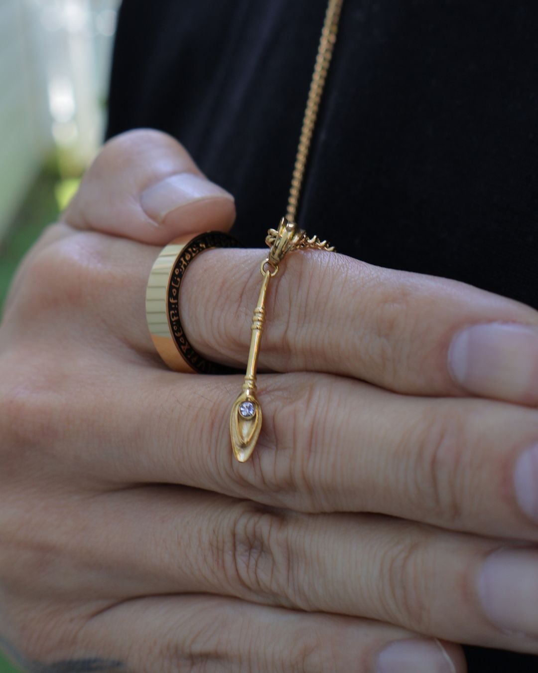 A gold pendant necklace with a stylized teardrop-shaped charm and a small clear gemstone, worn on a person's finger alongside other rings, set against a dark shirt background.