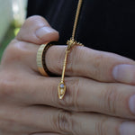 A gold pendant necklace with a stylized teardrop-shaped charm and a small clear gemstone, worn on a person's finger alongside other rings, set against a dark shirt background.