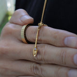 A gold pendant necklace with a stylized teardrop-shaped charm and a small clear gemstone, worn on a person's finger alongside other rings, set against a dark shirt background.