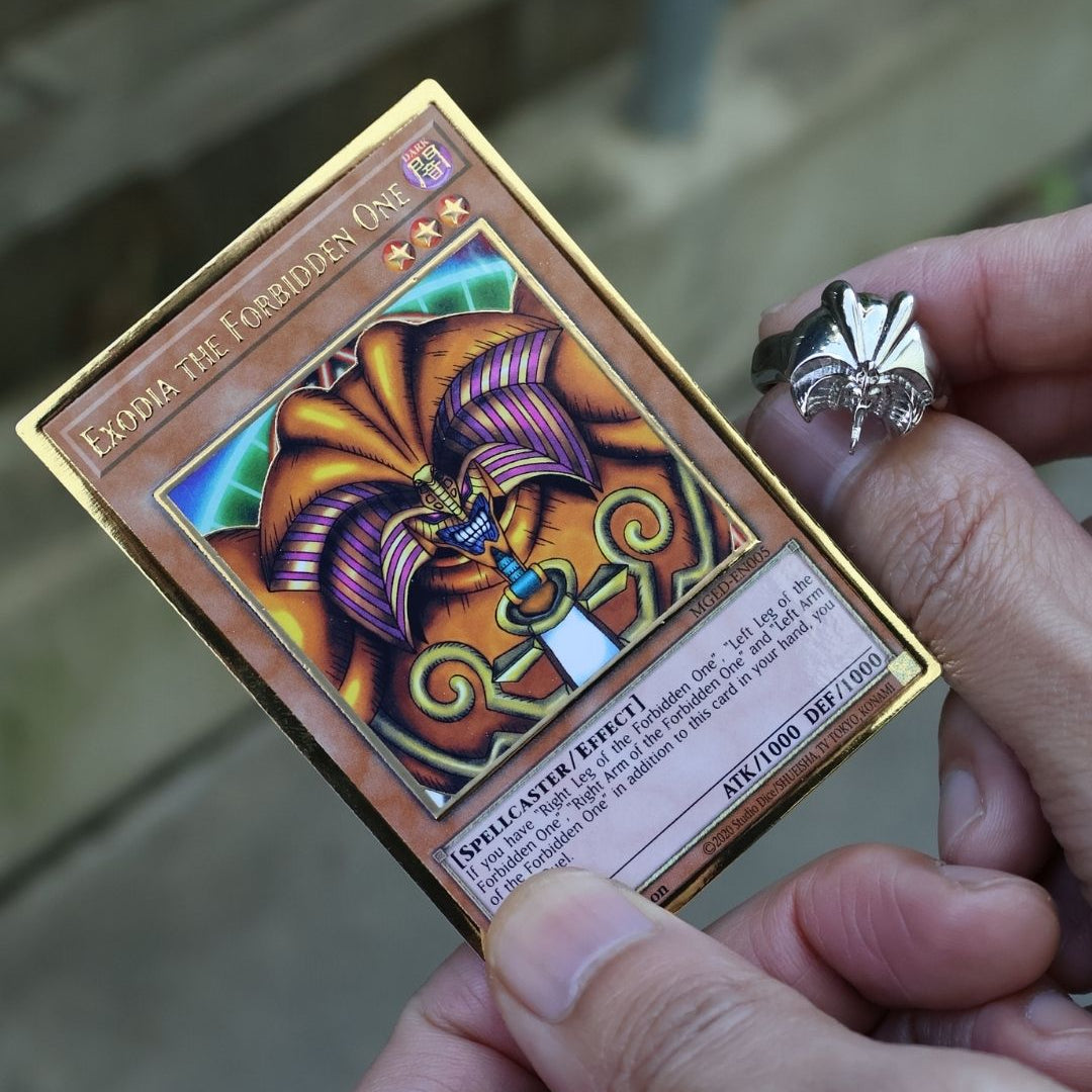 A close-up of a person holding a Yu-Gi-Oh! trading card, Exodia: The Coordinates One, with a silver ring on their finger, set against a blurred outdoor background.