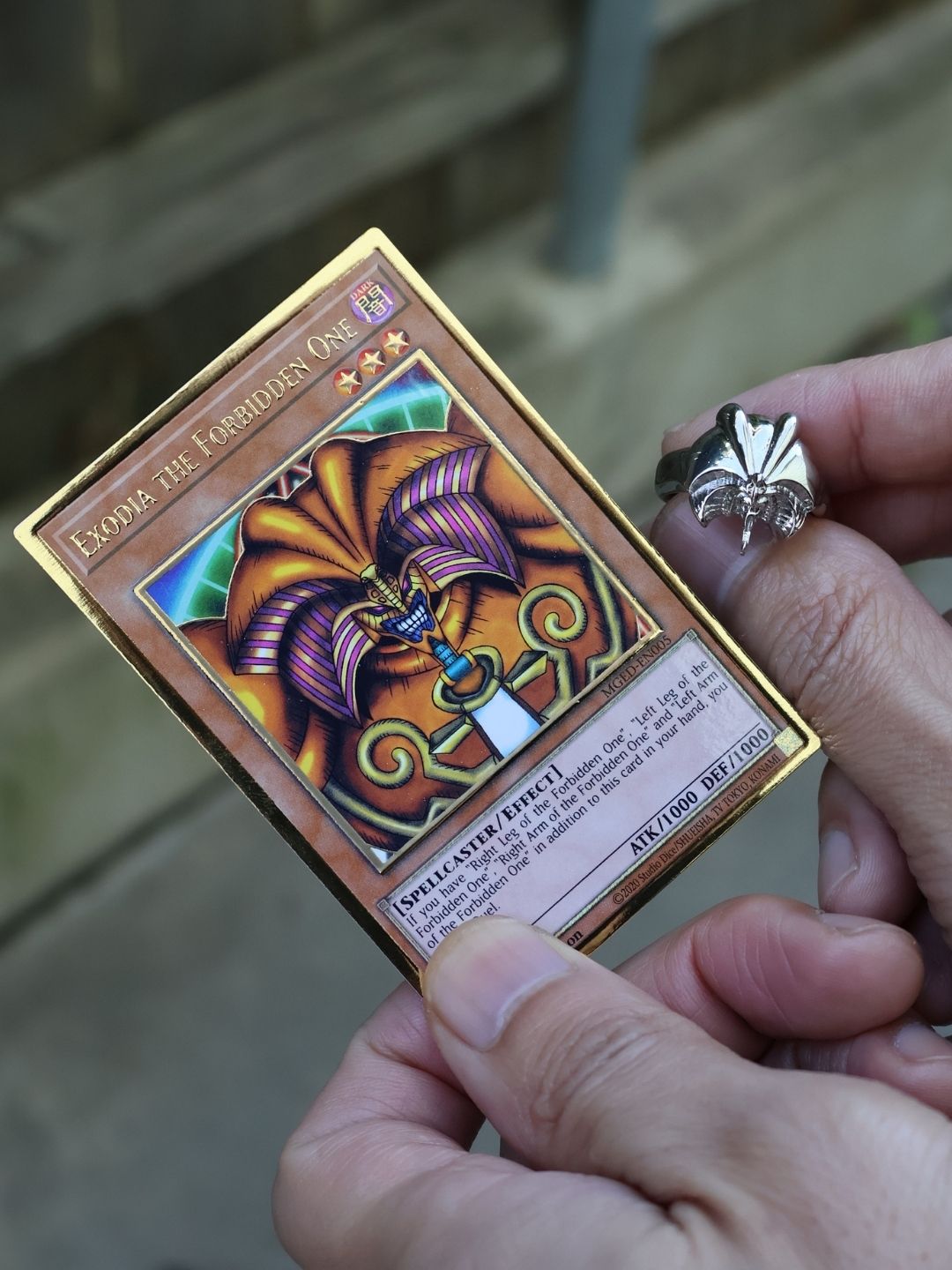 A close-up of a person holding a Yu-Gi-Oh! trading card, Exodia: The Coordinates One, with a silver ring on their finger, set against a blurred outdoor background.
