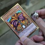 A close-up of a person holding a Yu-Gi-Oh! trading card, Exodia: The Coordinates One, with a silver ring on their finger, set against a blurred outdoor background.