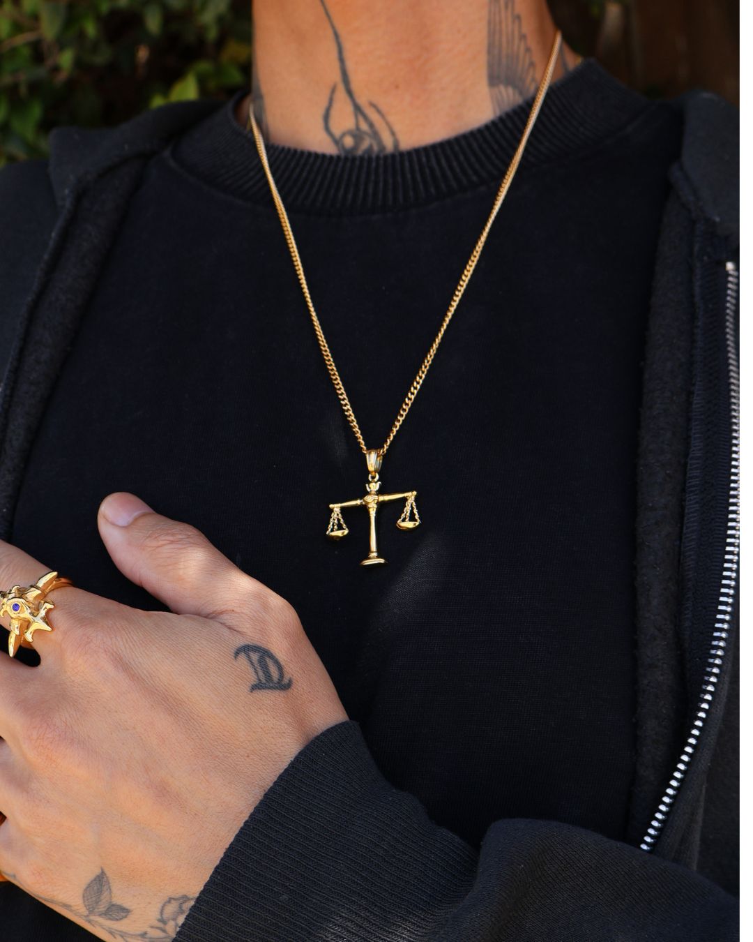 A gold chain necklace with a small balance scale pendant, worn by a person in a black zip-up hoodie. The person also has a gold ring on their hand.