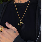 A gold chain necklace with a small balance scale pendant, worn by a person in a black zip-up hoodie. The person also has a gold ring on their hand.