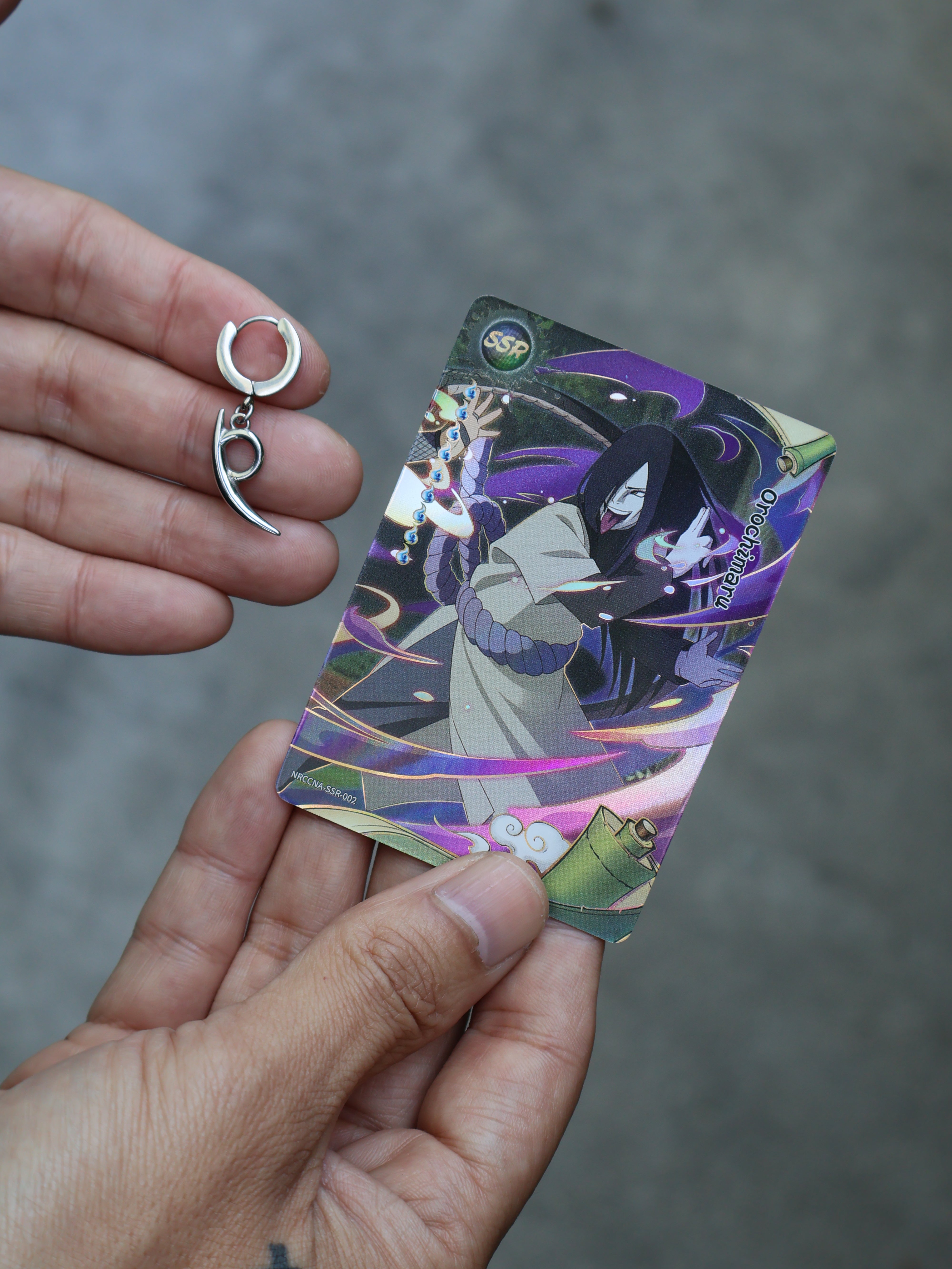 A person's hands holding a Naruto trading card featuring Sasuke Uchiha and holding a small silver crescent-shaped hoop earring next to it.