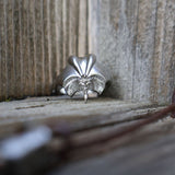 A silver ring with an intricate, sculptural design resembling a stylized flower or leaf cluster, photographed resting on a weathered wooden surface between two wooden planks.