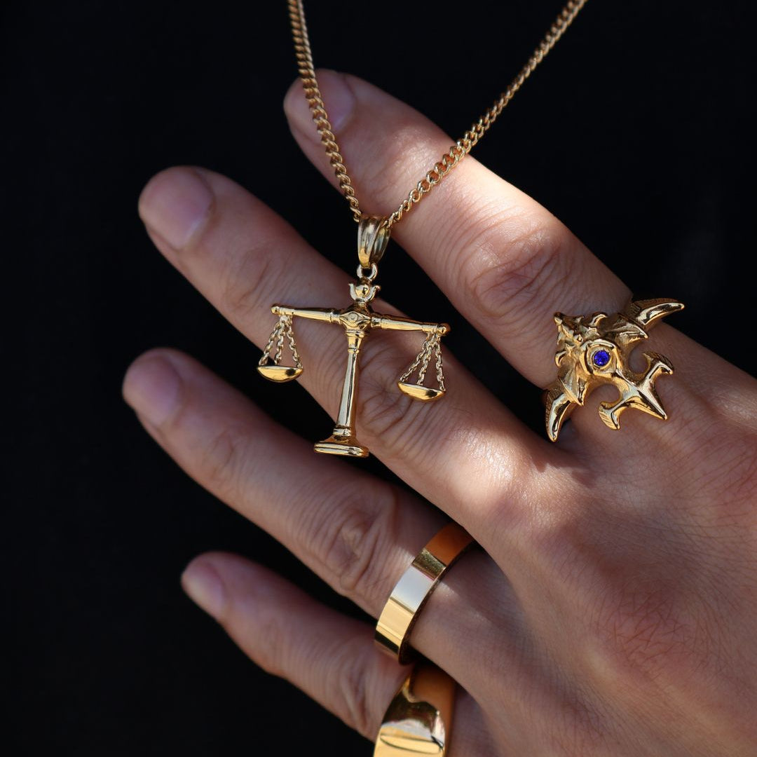 A gold-tone necklace with a scale of justice pendant is shown worn on a hand, alongside matching gold rings, against a solid black background.